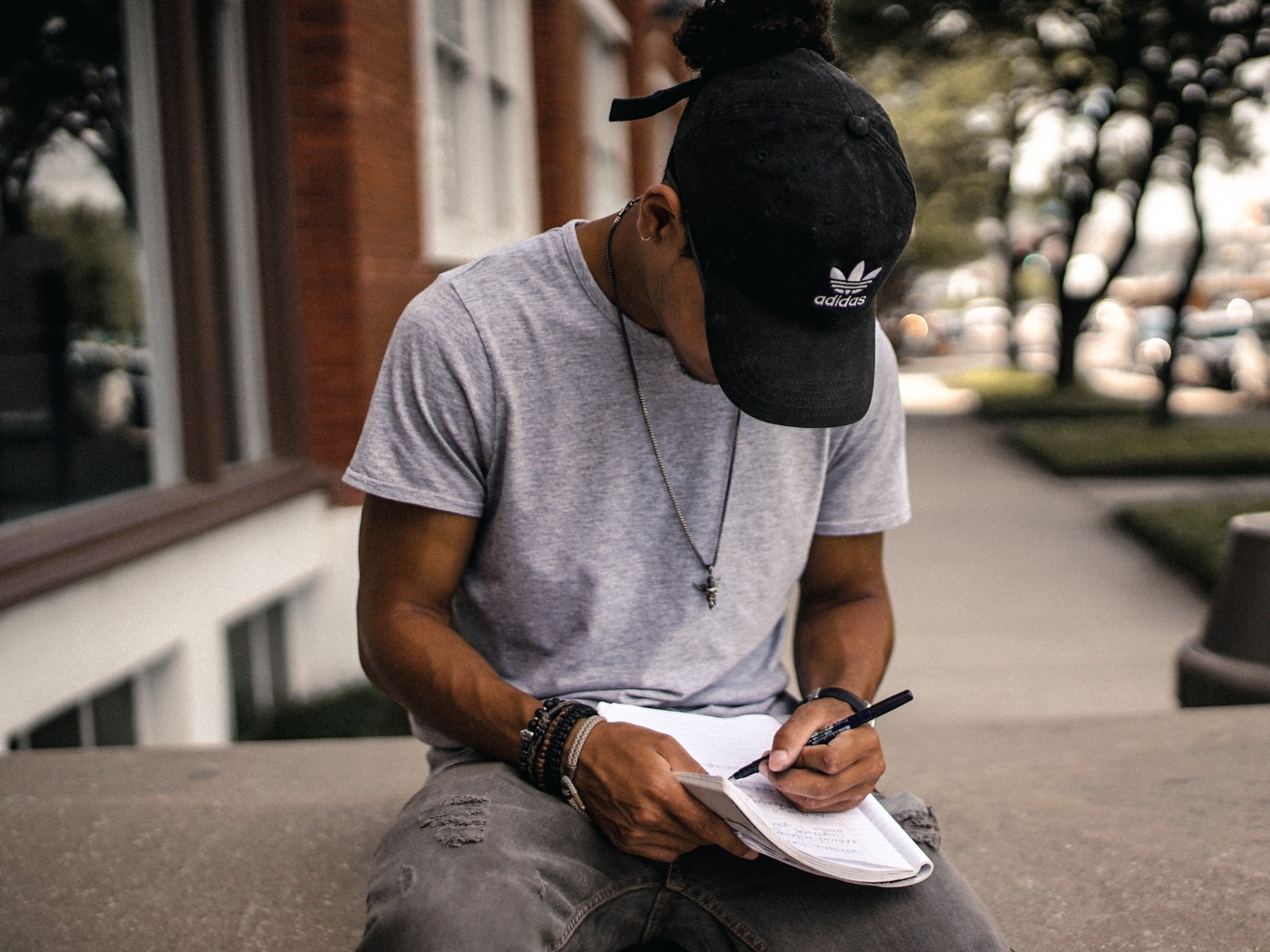 Young male seating on a step street-side, wearing a hat, head down writing in a journal