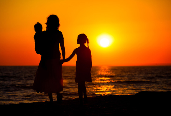 Shilluoette of woman hold a baby on her hip and holding hands with a young girl while walking on a beach at sunset
