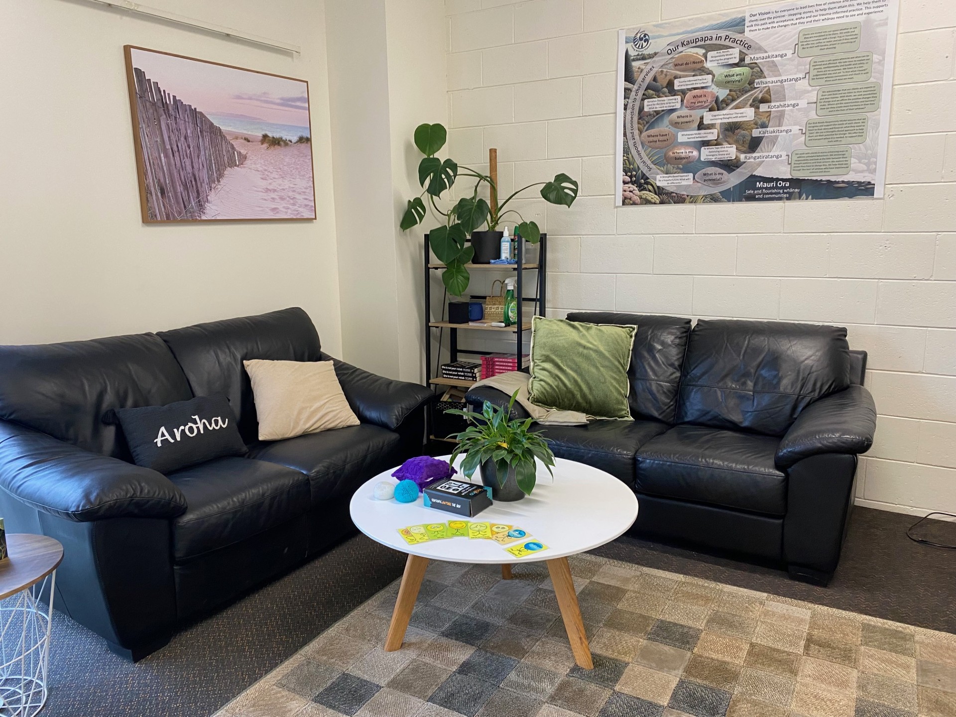 Welcoming comfy couch area in the Kapati Living Without Violence office. Pictures on the wall, plant on a coffee table, bookshelf in the background. Cushion with Aroha on it.
